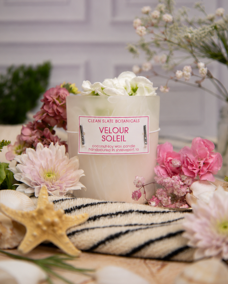 Candle labeled 'Velour Soleil' surrounded by flowers and seashells on a decorative surface.