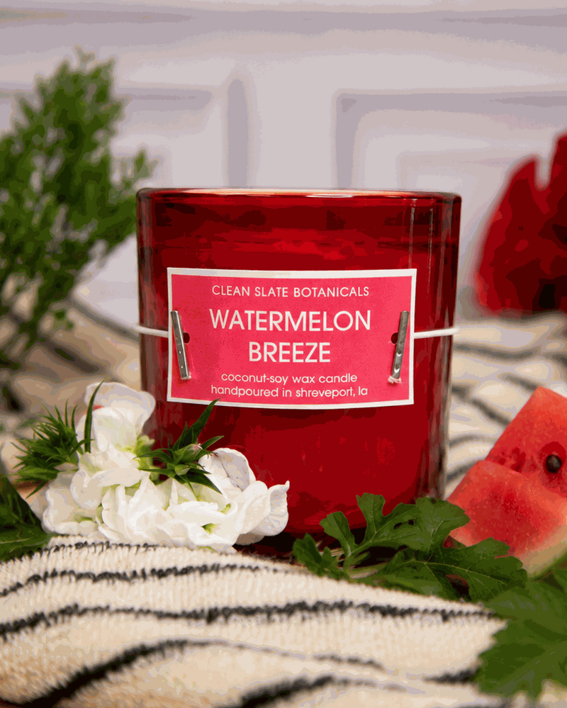 Watermelon Breeze Candle in a translucent red glass jar with a red label and white lettering, beside watermelon